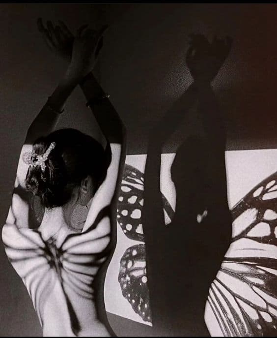 A black-and-white photograph of a person with their arms raised, featuring butterfly-wing patterns projected onto their back and a wall behind them. The image captures the interplay of light and shadow, creating a silhouette of the person's upper body and arms on the right side.