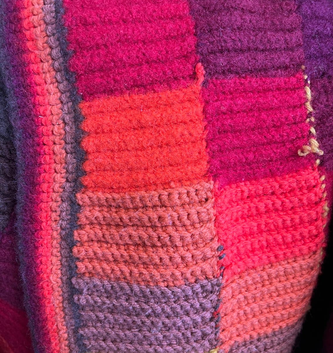 A close-up view of a hand-crocheted textile featuring a patchwork of rectangular blocks in warm tones of red, orange, and purple. The yarn has a fuzzy, felted texture, and the varying degrees of felting between the blocks create a slightly uneven, distorted surface.