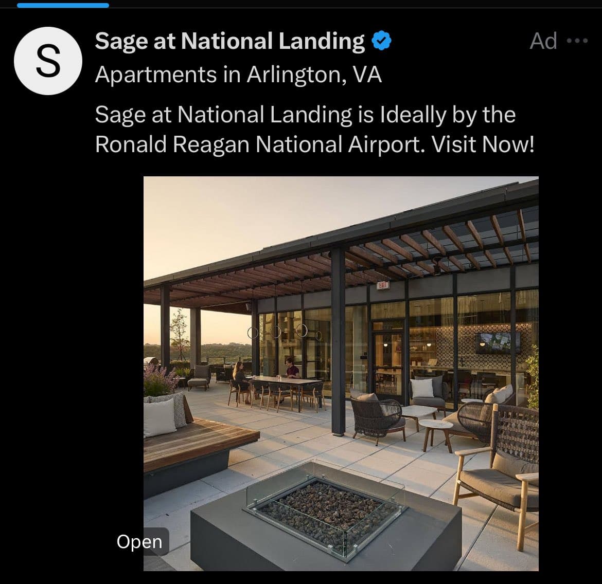 A screenshot of a Twitter ad from the verified account 'Sage at National Landing' promoting apartments in Arlington, VA. The ad features a photo of a modern outdoor patio area with lounge furniture, a fire pit, and a glass-walled building at dusk.