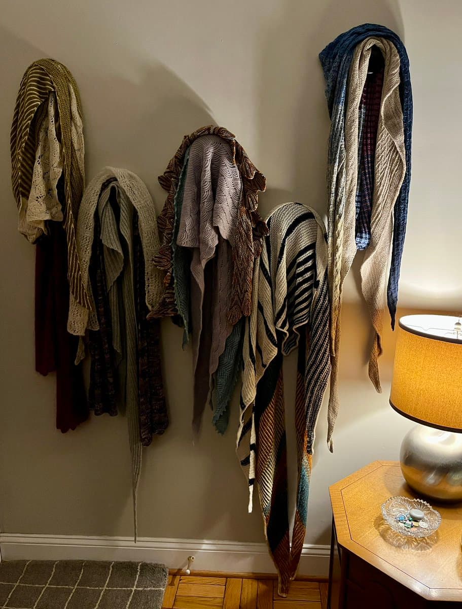 A collection of colorful, hand-knitted shawls and wraps is draped over hooks on a white wall, showcasing various patterns and textures. A lamp and a small table are visible in the lower right corner.