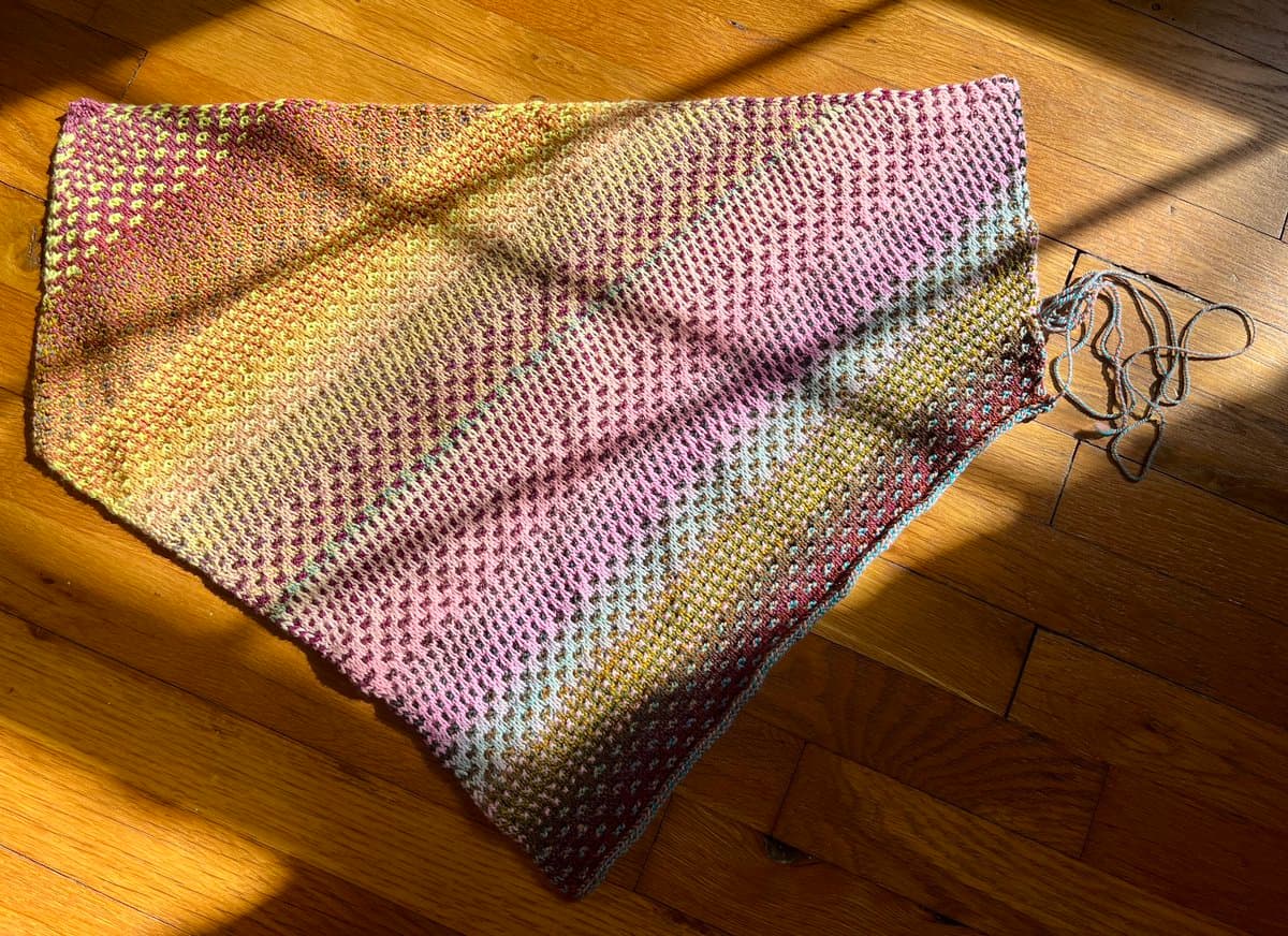 A flat, rectangular piece of multicolored knitted fabric lies on a wooden floor, illuminated by a diagonal beam of sunlight. A loose end of yarn is visible on the right side of the knitting.