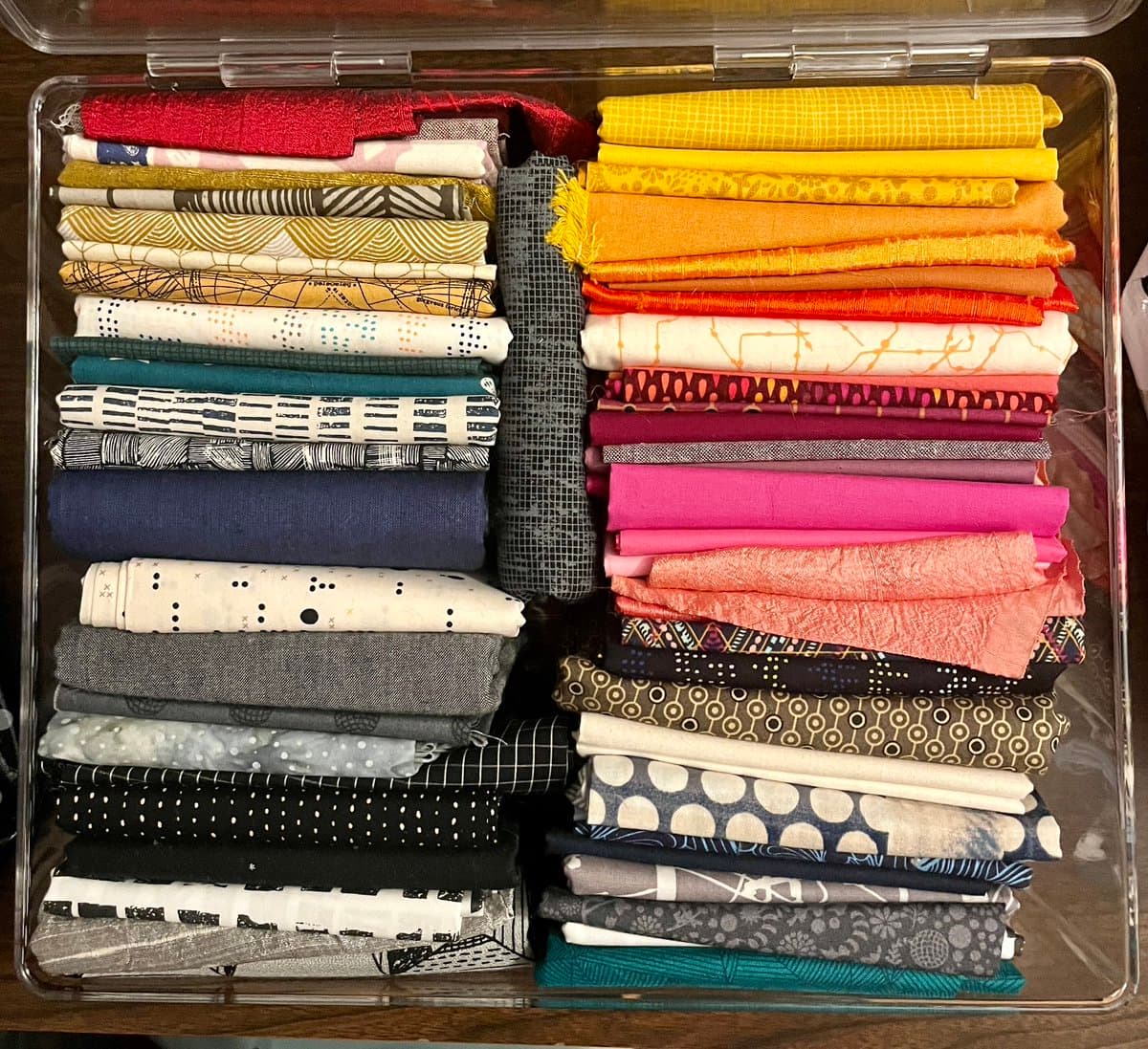 A top-down view of a clear plastic storage bin filled with neatly folded strips of fabric in various colors and patterns, organized into two columns. The fabrics feature a wide range of hues including reds, yellows, oranges, pinks, blues, and grays, with designs ranging from solid colors to polka dots, stripes, and geometric prints.