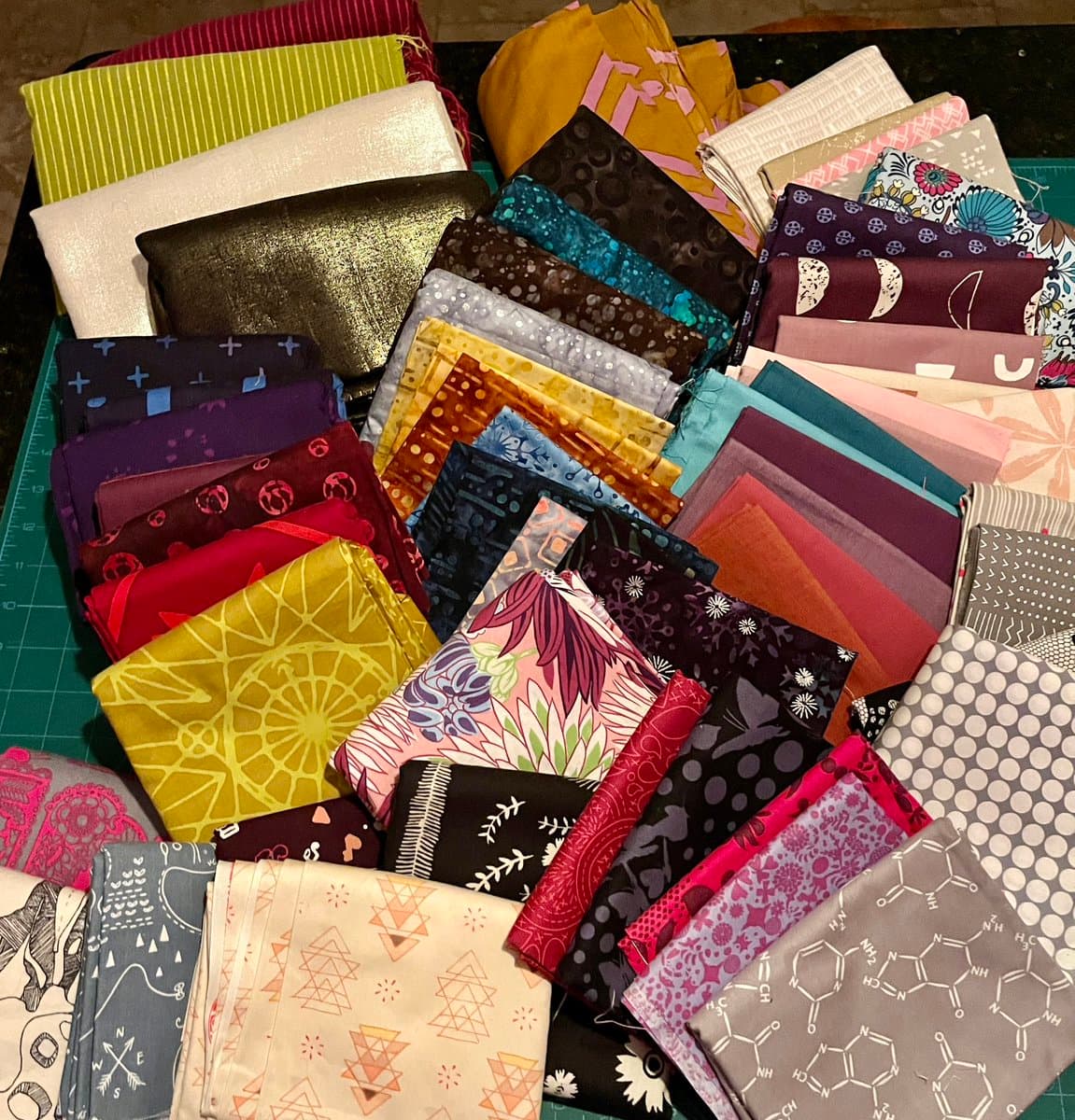 A top-down view of numerous folded pieces of colorful, patterned fabric arranged in a pile on a green cutting mat. The fabrics feature various designs, including geometric shapes, floral prints, and abstract patterns in a wide range of colors.