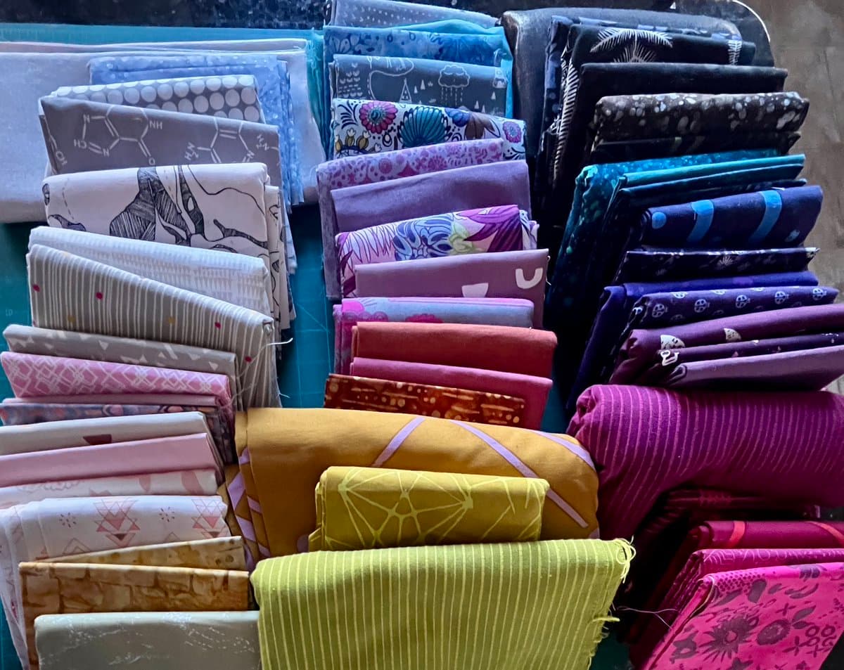 A collection of folded fabric pieces in various colors and patterns, including floral, geometric, and abstract designs, arranged in stacks. The fabrics appear to be pre-cut squares or rectangles, likely intended for quilting or crafting.