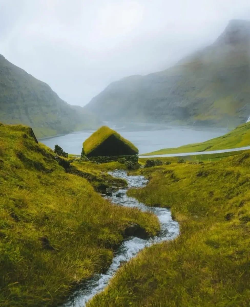 A landscape photograph of a misty mountain valley featuring a small, grass-roofed hut near a body of water, with a stream flowing through the green foreground.