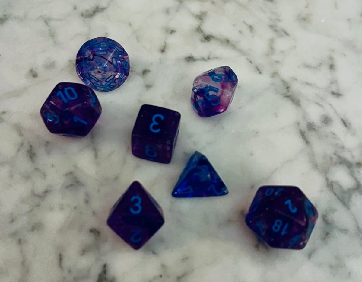 A photograph of a set of seven purple and blue polyhedral dice scattered on a white marble surface.
