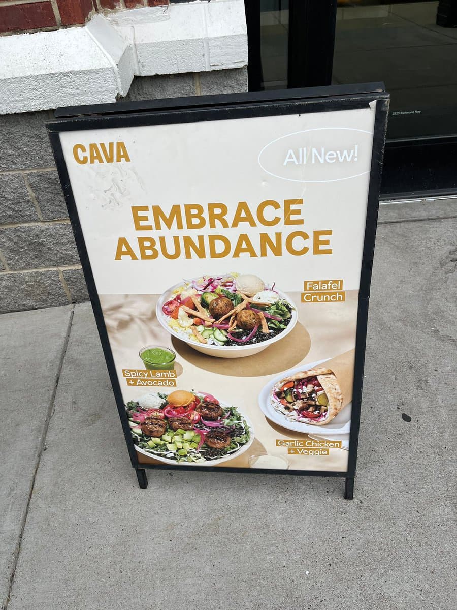A sidewalk advertising sign for the restaurant CAVA, featuring the slogan "Embrace Abundance" and images of three new menu items: a Falafel Crunch bowl, a Spicy Lamb + Avocado bowl, and a Garlic Chicken + Veggie wrap.