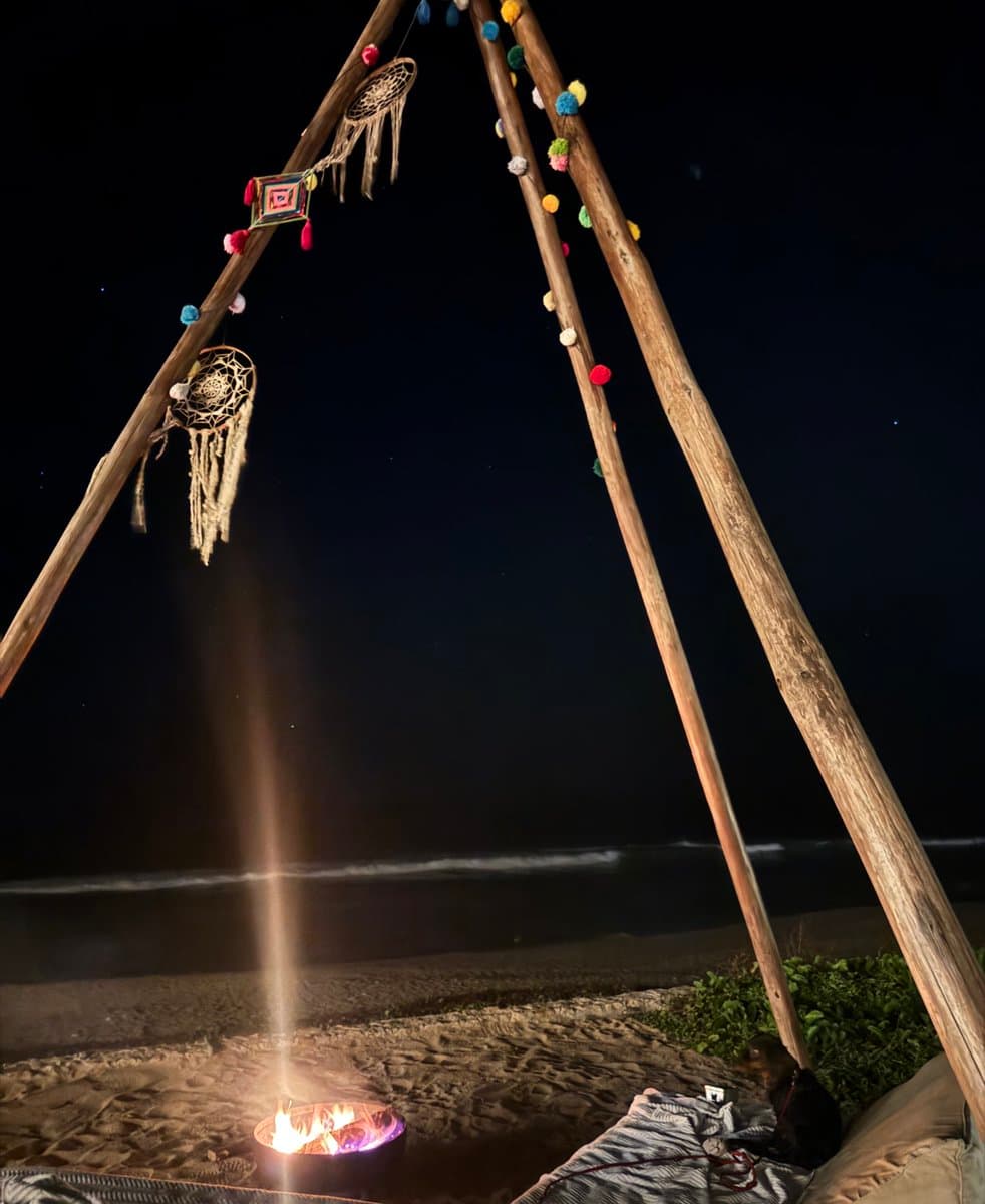 A nighttime photograph of a beach campsite featuring a small fire burning in a metal fire pit, framed by wooden poles decorated with dreamcatchers and colorful pom-poms, with the ocean and starry sky in the background.