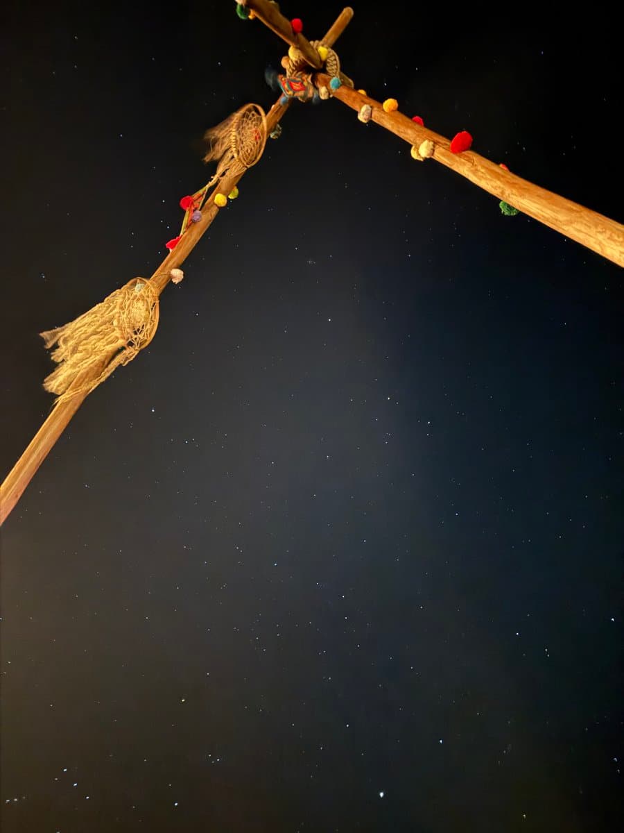 A photograph of a decorated wooden dreamcatcher frame silhouetted against a dark night sky filled with stars. The frame is adorned with colorful beads and woven webbing, with the stars in the background resembling the freckles mentioned in the caption.
