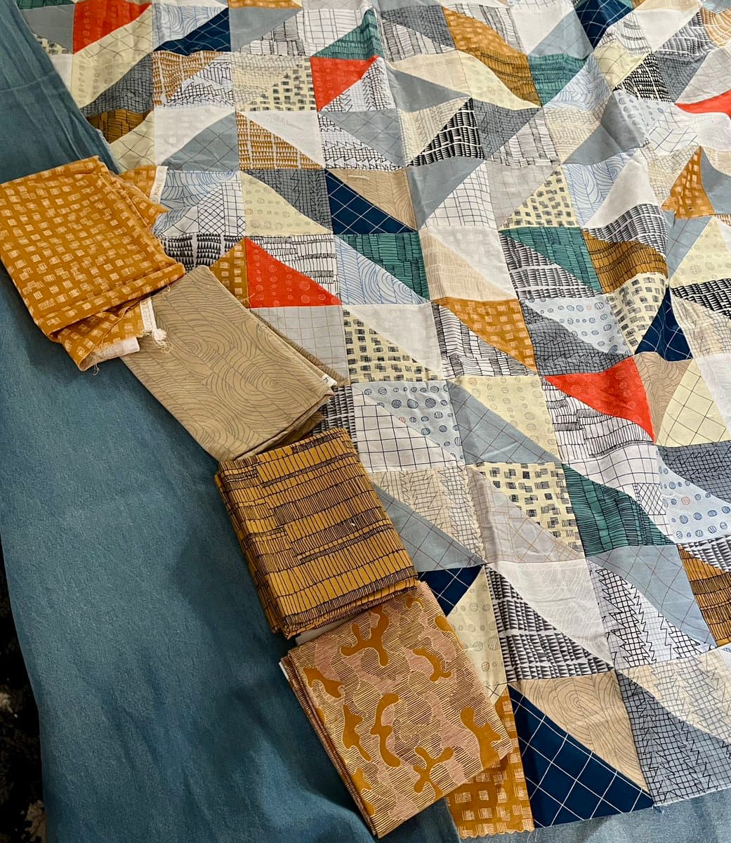 A top-down view of a colorful patchwork quilt featuring geometric triangle patterns in shades of blue, orange, yellow, and white, laid out on a blue surface. Several folded pieces of patterned fabric are arranged in a row along the left edge of the quilt.