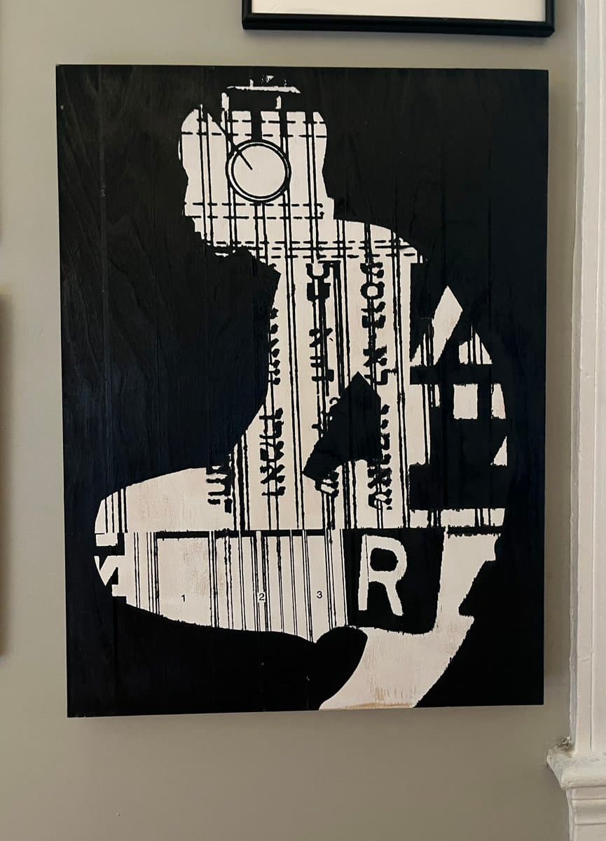 A black and white painting on a wooden canvas depicting a silhouette of a person playing a guitar, with the guitar body filled with musical notation and text. The artwork hangs on a light-colored wall next to a white door frame.