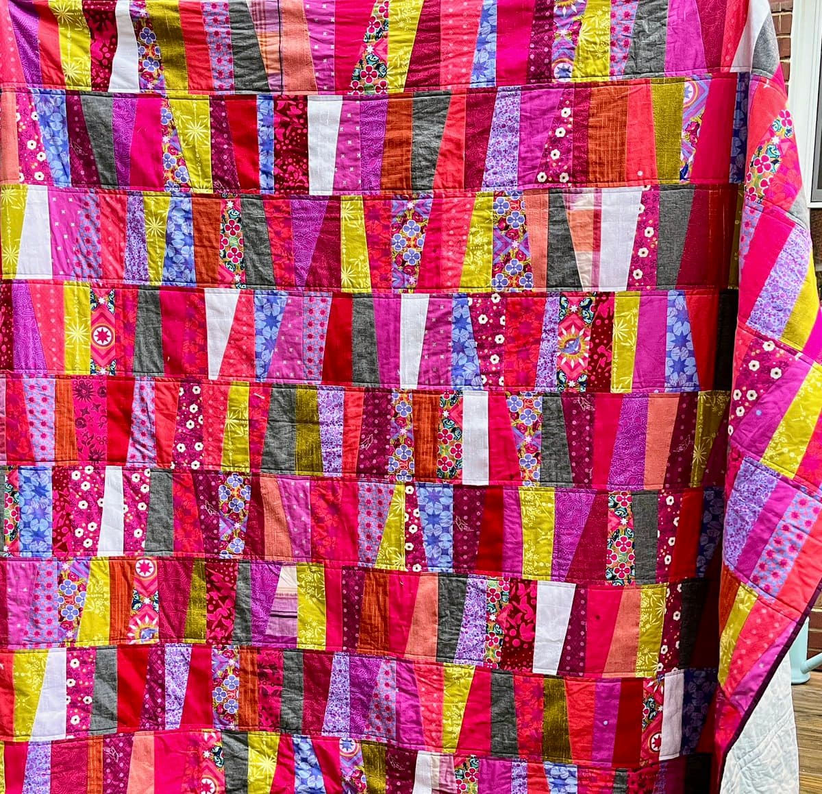 A close-up view of a colorful patchwork quilt featuring a geometric pattern of vertical strips in shades of pink, red, purple, yellow, and blue. The fabric pieces have various floral and abstract prints, and the quilt is draped over a surface, with a white pillow visible in the bottom right corner.