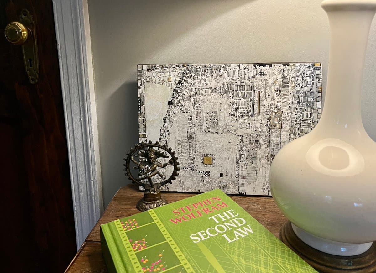 A still-life photograph of a wooden table featuring a copy of Stephen Wolfram's book 'The Second Law', a small metallic figurine of a figure on a gear, a white ceramic vase, and a framed piece of abstract, mosaic-style art in the background.