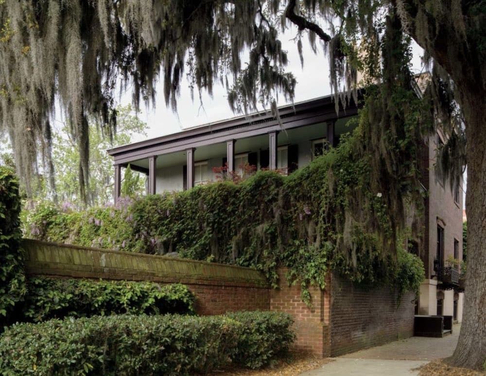 A historic brick house in Savannah, Georgia, is framed by large oak trees draped in Spanish moss, with a lush, ivy-covered brick wall in the foreground. The scene captures the city's characteristic Southern charm and natural beauty.