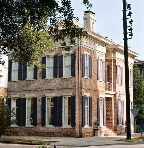 A two-story brick building with white trim and dark shutters, featuring a corner entrance and a chimney on the roof. The structure is framed by trees and a streetlamp, set against a clear sky.