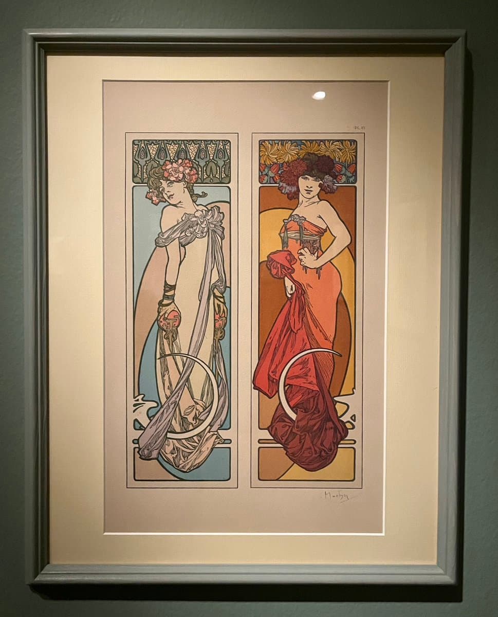 A framed Art Nouveau print by Alphonse Mucha, displayed in a gallery, featuring two stylized female figures in flowing dresses within vertical panels. The artwork is signed by the artist in the lower right corner.