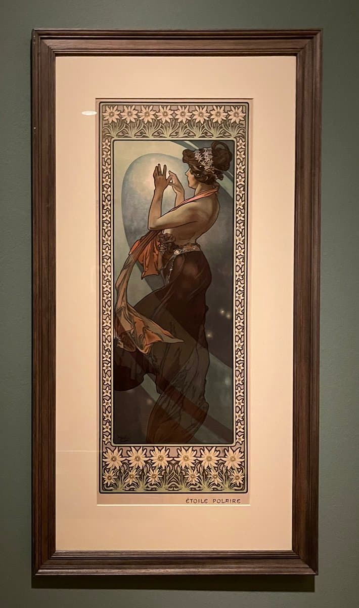 A framed Art Nouveau lithograph by Alphonse Mucha, depicting a woman in profile reaching toward a large moon, set within an ornate floral border. The piece is displayed in a dark wooden frame with a cream mat against a gray wall. The title 'ÉTOILE POLAIRE' is printed at the bottom of the artwork.
