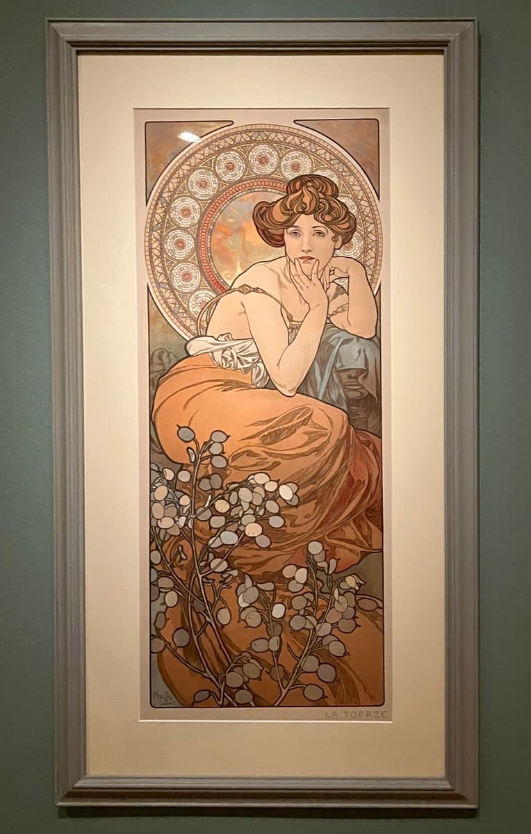 A framed Art Nouveau lithograph by Alphonse Mucha, titled 'La Topaze,' depicting a woman in an orange dress seated against a decorative circular background. The artwork is displayed in a gallery setting, likely at the Phillips Collection.