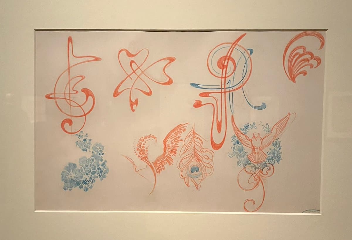 A framed piece of decorative art featuring stylized, ornamental motifs in orange and blue ink on a white background. The design includes abstract flourishes, floral clusters, and a bird-like figure, characteristic of Art Nouveau style.