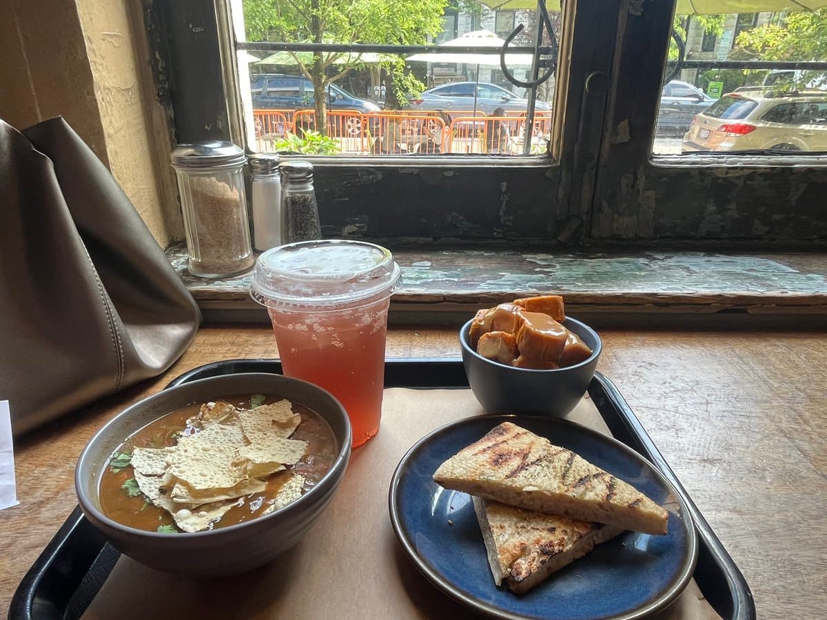 A meal served on a black tray at a window-side table, featuring a bowl of soup with tortilla chips, a plate of grilled sandwich halves, a small bowl of fried snacks, and a plastic cup of pink iced drink. Through the window, a street scene with parked cars and outdoor cafe umbrellas is visible.