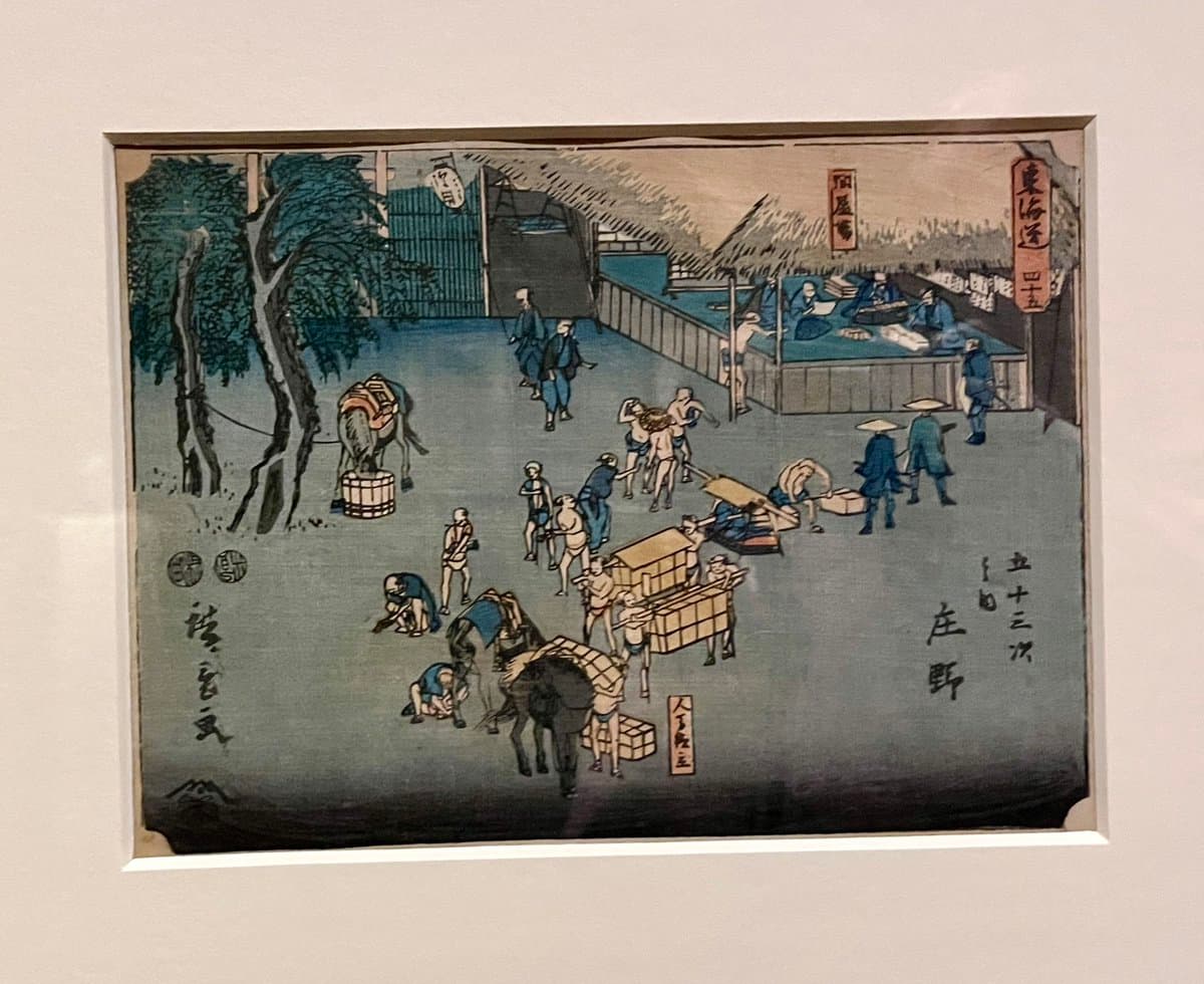 A framed Japanese woodblock print depicting a busy scene of travelers and porters with horses and carts at a station, featuring Japanese text in the corners.