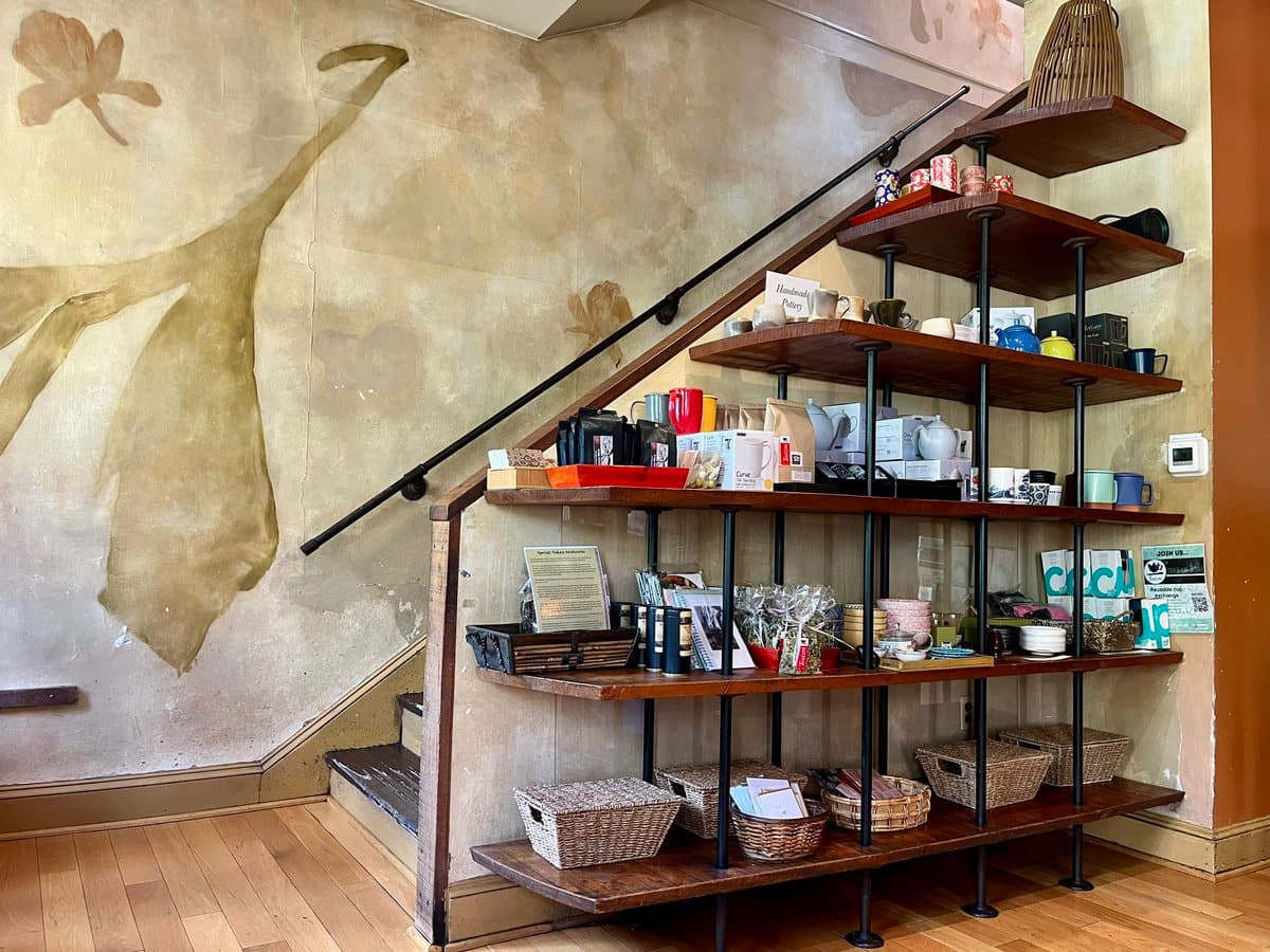 A photograph of a wooden, multi-tiered display shelf built into the side of a staircase, holding various mugs, teapots, and baskets. The wall behind the shelves features a large, faded mural of a figure in a dress.