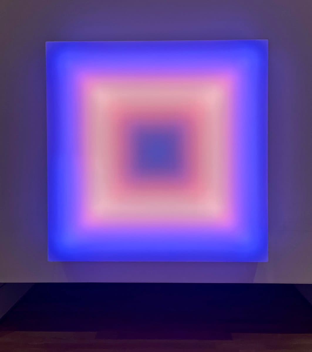 A photograph of a large, square abstract artwork hanging on a gallery wall. The piece features a glowing, blurred gradient that transitions from a deep blue outer border to a soft pink and white center, creating a tunnel-like visual effect.