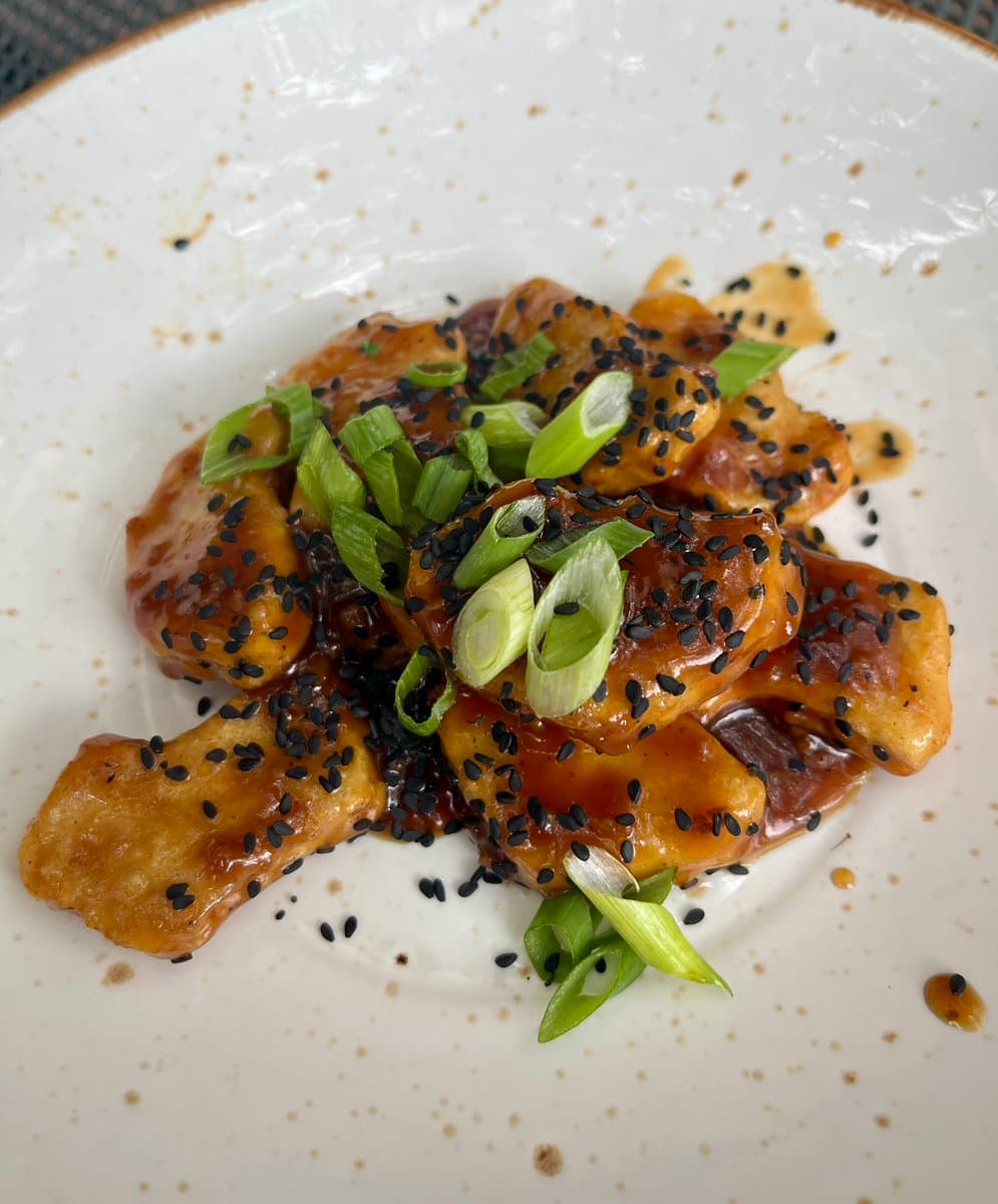 A close-up, top-down shot of a plate of vegan nuggies coated in a glossy brown sauce, garnished with black sesame seeds and sliced green onions.