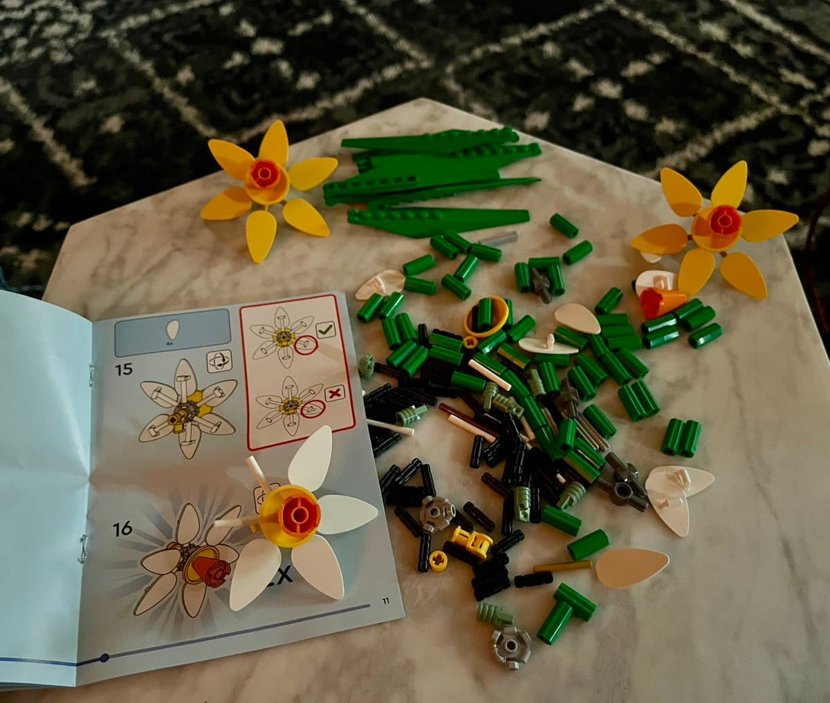 A top-down view of a LEGO build in progress on a marble table, featuring two completed yellow daffodil flowers, a pile of green and black LEGO pieces, and an open instruction manual. The manual displays step-by-step diagrams for assembling the flowers, with steps 15 and 16 visible.