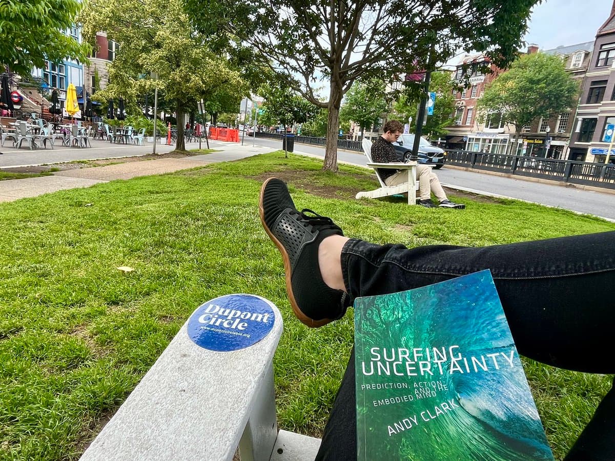 A first-person perspective photo of a person relaxing on a park bench, with their legs crossed and a book resting on their lap. In the background, a street scene includes trees, a cafe, and another person sitting on a bench. The book's cover is clearly visible, as is a sticker on the bench.