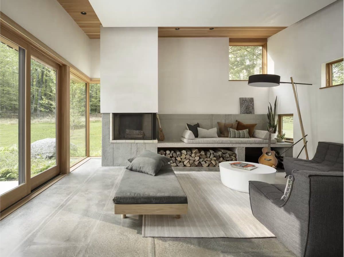 A photograph of a modern, minimalist living room featuring a concrete fireplace, built-in concrete bench seating, and large glass doors that open to a grassy, wooded exterior. The space is furnished with a gray chaise lounge, a gray sofa, a white round coffee table, and a floor lamp, with a guitar leaning against the bench.