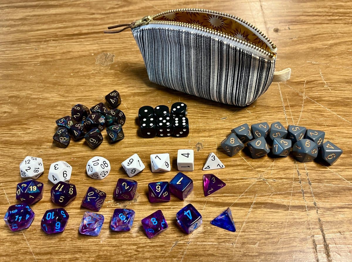 A collection of various tabletop role-playing game dice, including polyhedral and standard six-sided dice, arranged on a wooden table in front of an open striped dice pouch.