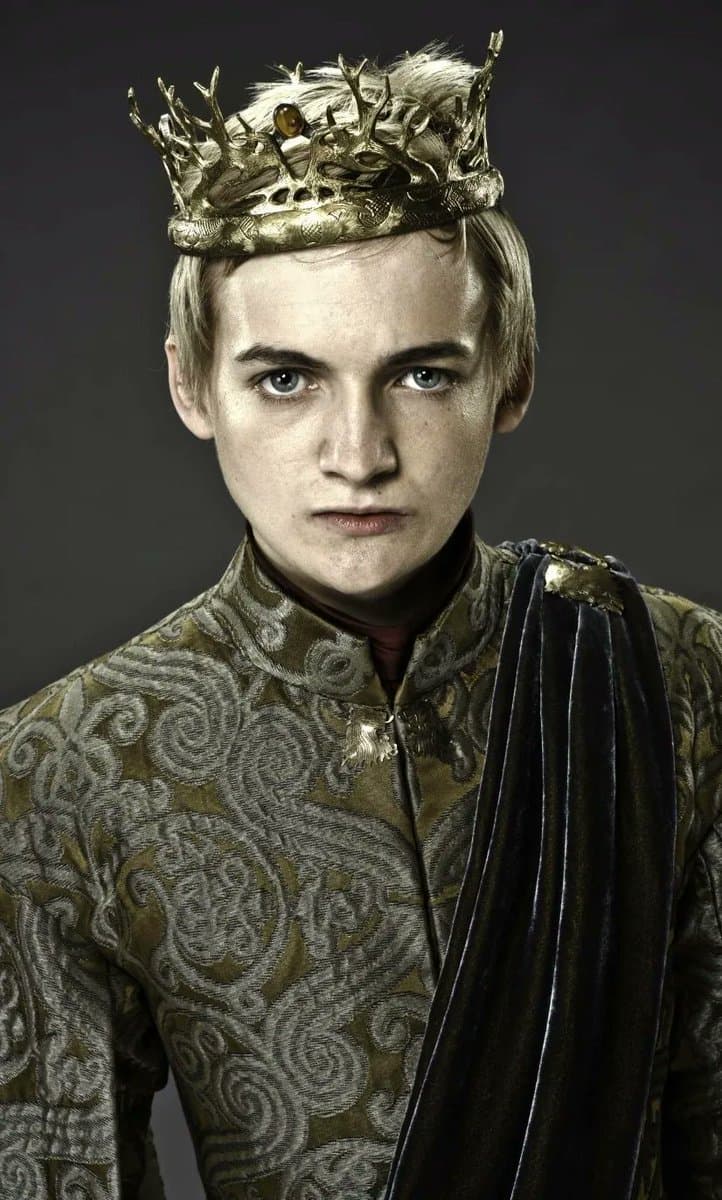A portrait of a young man with pale skin and light hair, wearing an ornate golden crown and a patterned green tunic with a dark velvet cloak draped over one shoulder. The image is a promotional still or character portrait, likely from a fantasy television series or film.
