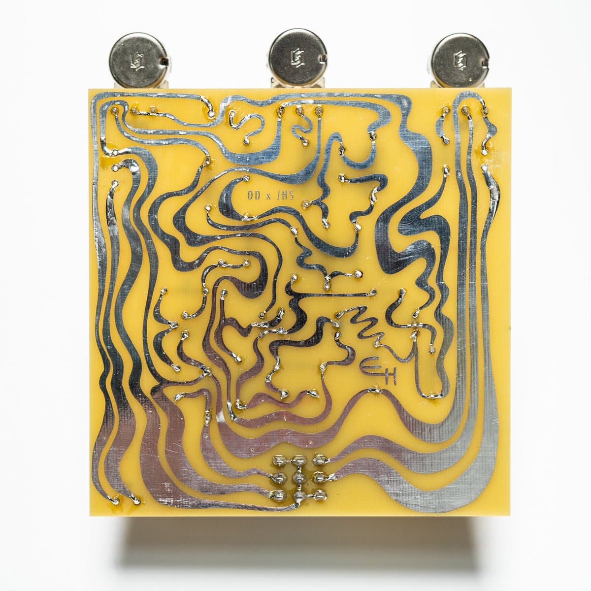 A close-up photograph of a yellow printed circuit board featuring intricate, wavy silver solder traces and three circular metal components at the top.