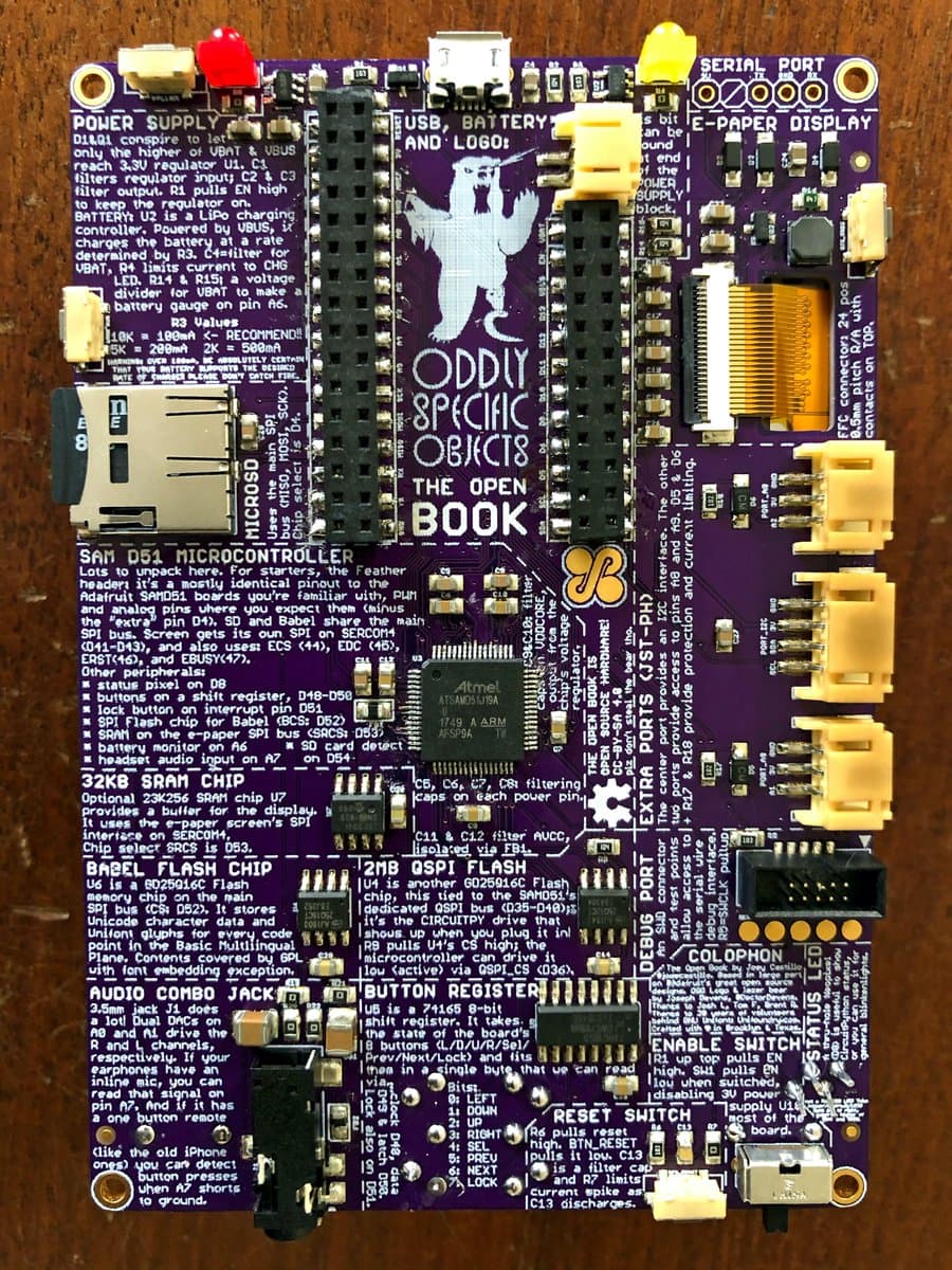 A close-up photograph of a purple printed circuit board (PCB) labeled 'Oddly Specific Objects The Open Book,' featuring a central microcontroller, various electronic components, and a white logo of a figure holding a book. The board is densely populated with text annotations explaining its different sections, such as power supply, microcontroller, and display interfaces.