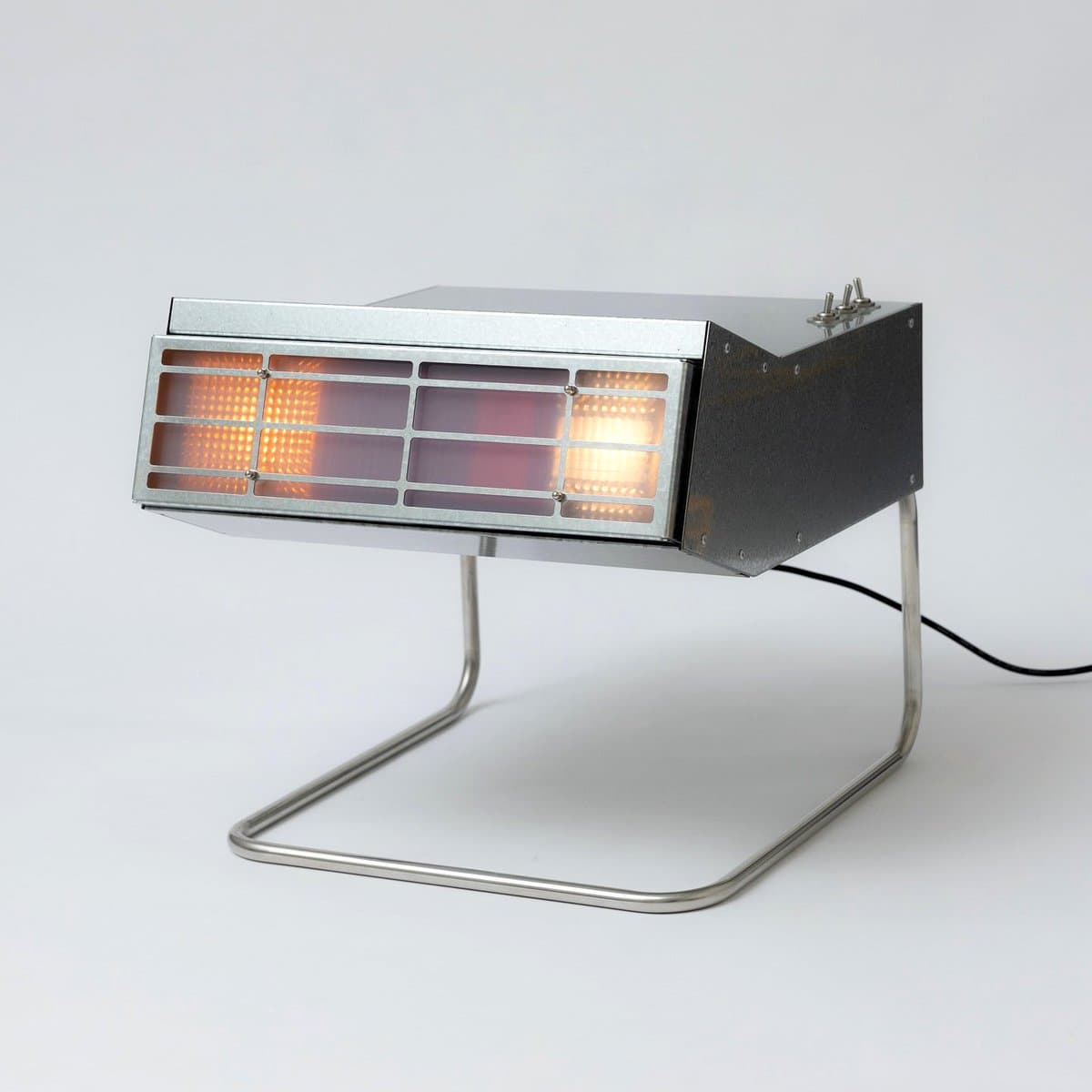 A product photograph of a modern, industrial-style steel lamp or side table. The object features a rectangular metal housing with a grid of illuminated, amber-colored panels on the front, supported by a thin, bent metal wire base.