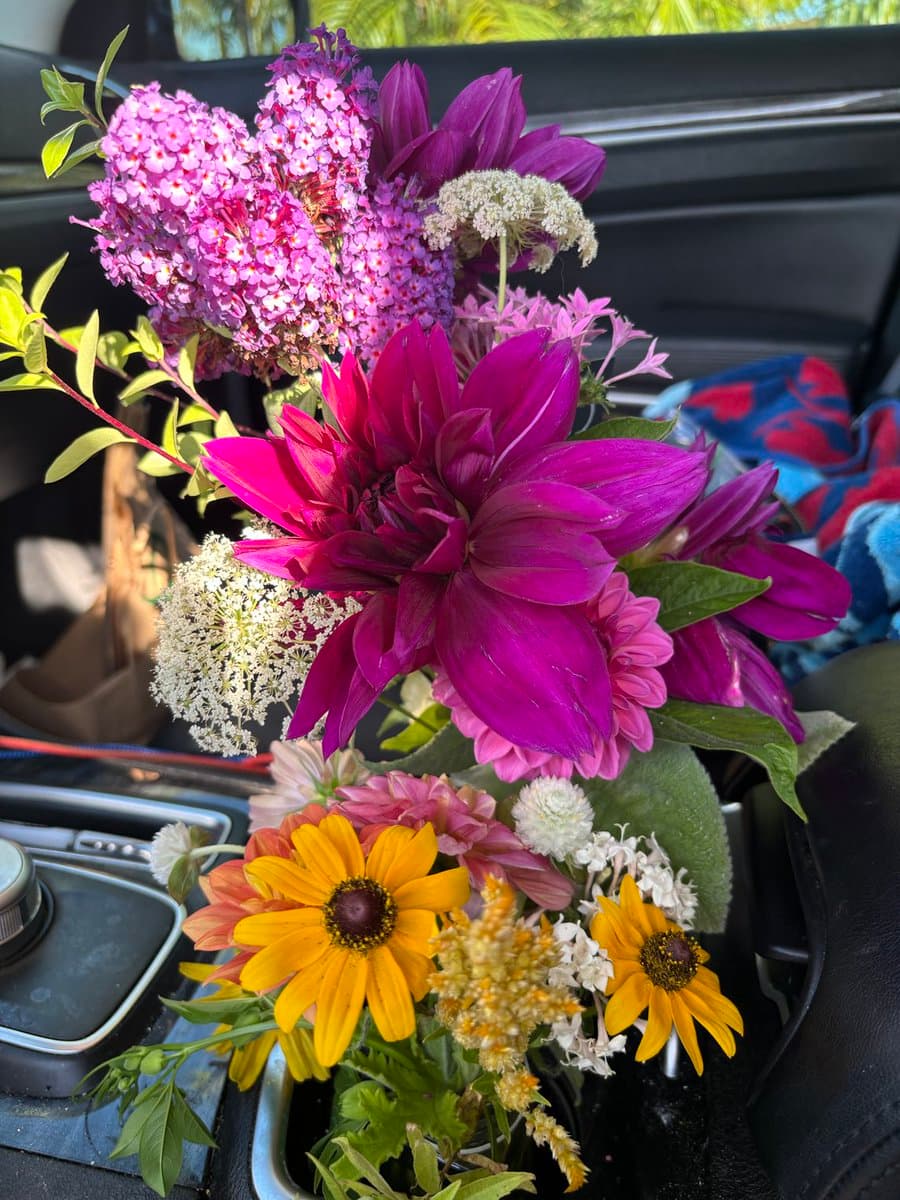 A vibrant bouquet of fresh flowers, including purple dahlias, pink blooms, and yellow sunflowers, is arranged in a container on the center console of a car.