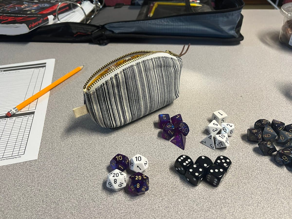A tabletop scene featuring multiple sets of polyhedral dice in purple, white, black, and gray, arranged next to a striped pencil case, a yellow pencil, and a character sheet on a gray surface.