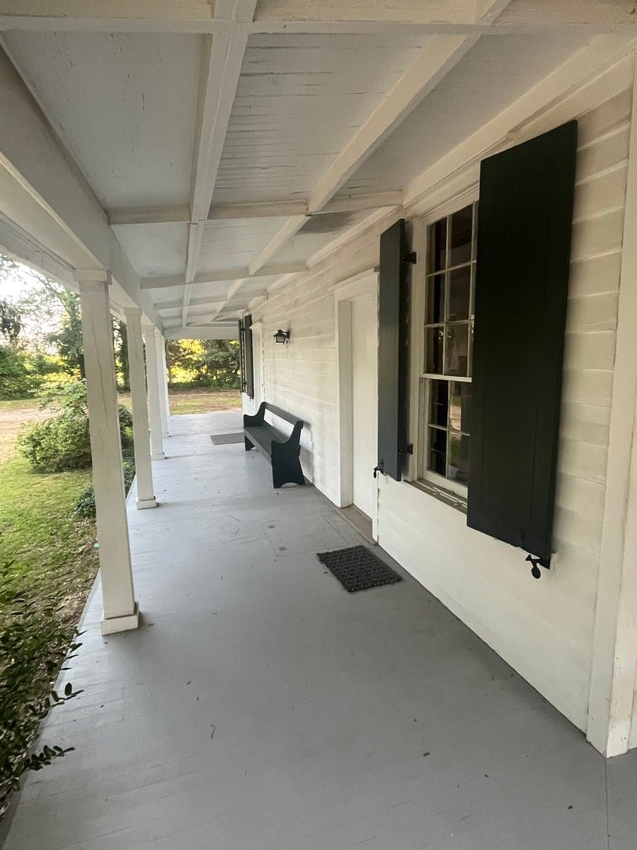 A long, covered porch of a white wooden building with black shutters, featuring a black bench and a doormat on the floor.