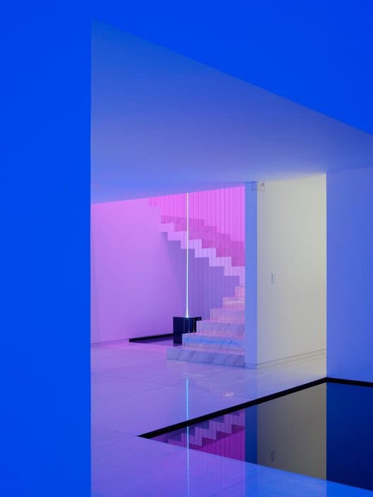 A minimalist interior space in a home, featuring a white staircase illuminated by vibrant pink and blue lighting. The scene includes a reflective pool in the foreground and a sleek, modern design with clean lines and smooth surfaces.