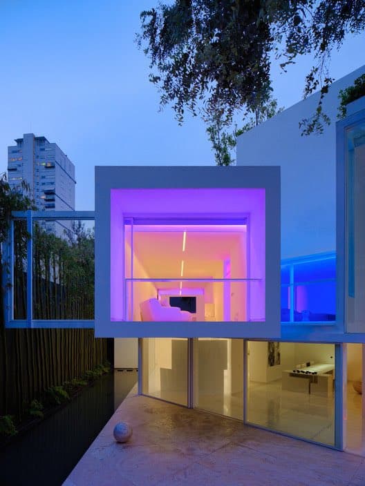 A minimalist white modern home in Mexico City is shown at dusk, featuring a cantilevered upper-level room illuminated with vibrant purple and orange lighting. The structure has large glass windows and a clean, geometric design, set against a twilight sky with a city building visible in the background.