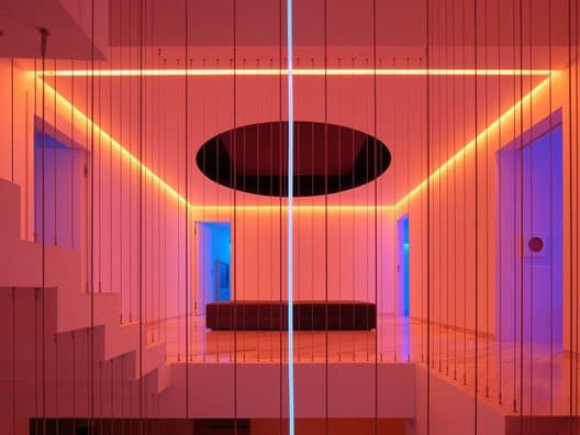 A minimalist interior space in a home, illuminated with vibrant pink and orange LED lighting that outlines the ceiling and walls. The room features a modern design with a circular ceiling fixture, a low bench, and a staircase on the left, creating a futuristic and artistic atmosphere.