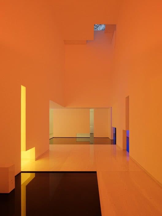 A minimalist interior space in a home, featuring smooth orange walls, a light wood floor, and a dark reflective pool or floor section. The room is illuminated by natural light filtering through a small skylight and colored light accents in yellow and blue.