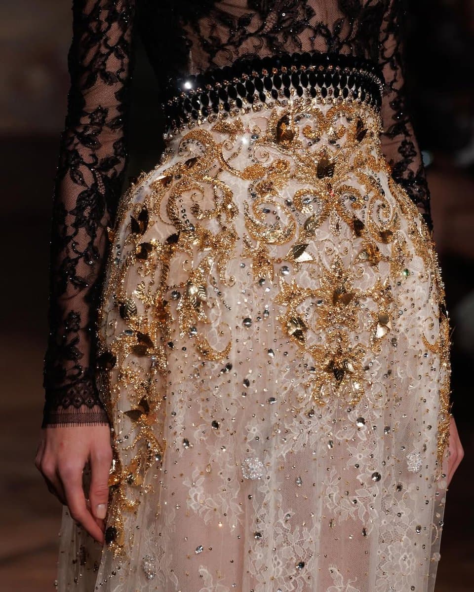 A close-up photograph of a model's torso and skirt, showcasing a black lace bodice and a sheer, ivory-colored skirt heavily embellished with intricate gold embroidery and crystal beading.