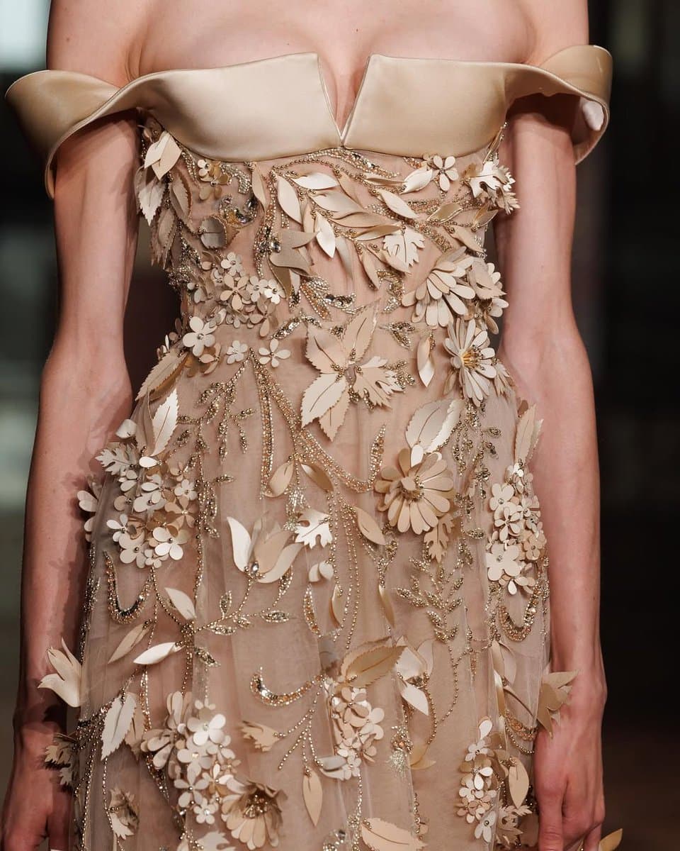 A close-up photograph of a model wearing an off-the-shoulder, beige couture gown featuring intricate 3D floral and leaf appliqués and beaded embroidery.