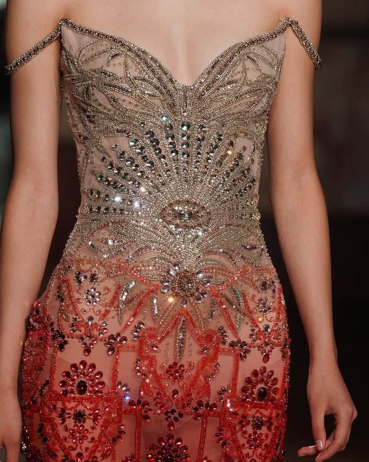A close-up photograph of a model wearing an intricately beaded and embroidered couture gown featuring a sheer bodice with silver embellishments and a skirt with red floral patterns.