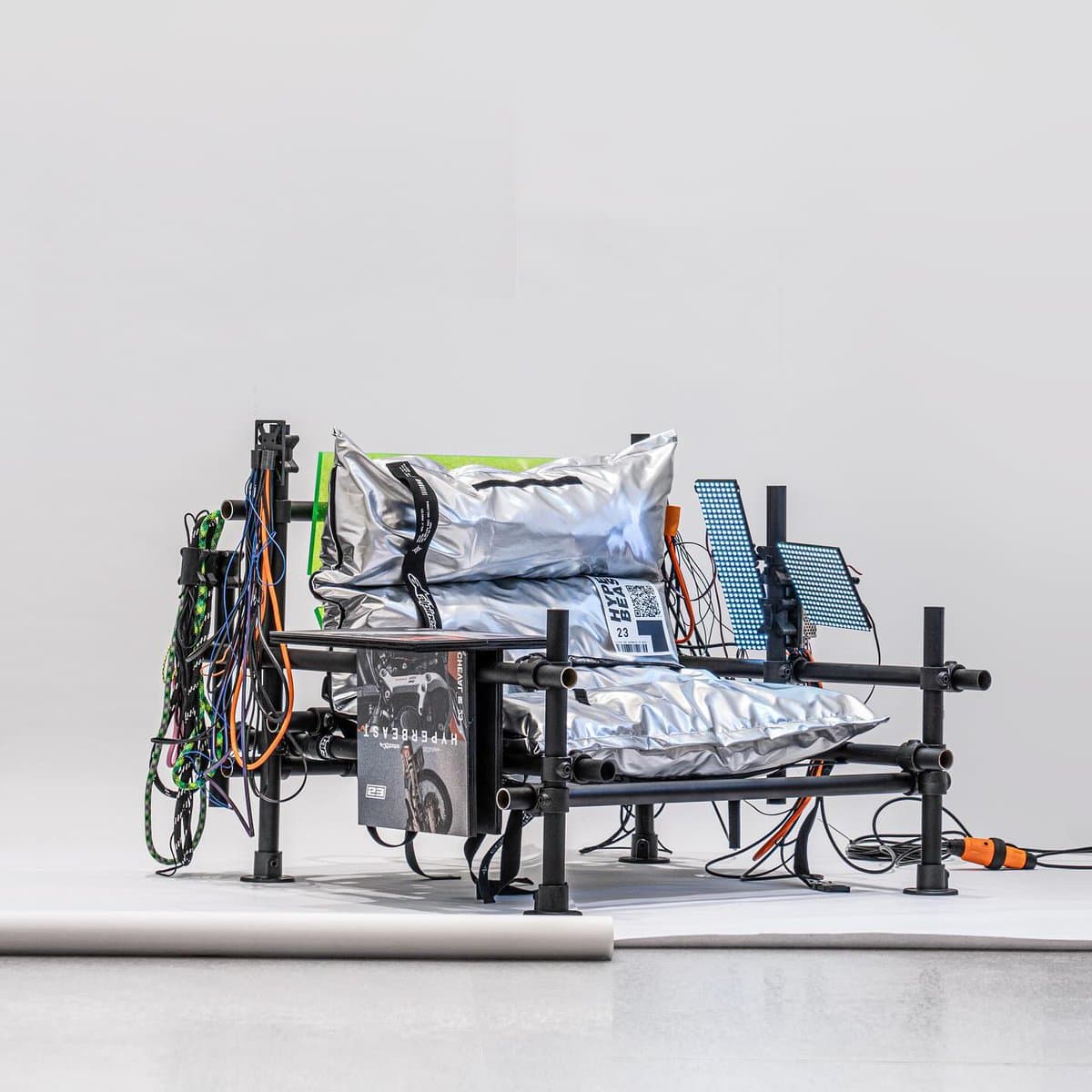 A studio photograph of a modern, industrial-style lounge chair constructed from black PVC pipes, featuring silver metallic cushions, hanging LED light strips, and bundles of colorful cables. A magazine titled 'HYPERBEAST' is placed on the armrest.