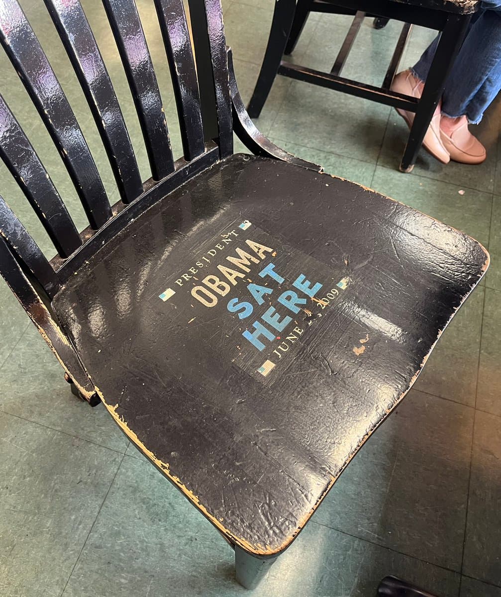 A close-up, angled shot of a worn black wooden chair with a sticker on its seat. In the background, the legs and feet of a person sitting on another chair are visible on a green tiled floor.