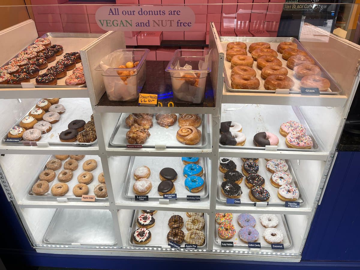 A display case filled with various types of donuts, including glazed, frosted, and filled varieties, arranged on multiple shelves. A sign above the case reads "All our donuts are VEGAN and NUT free."