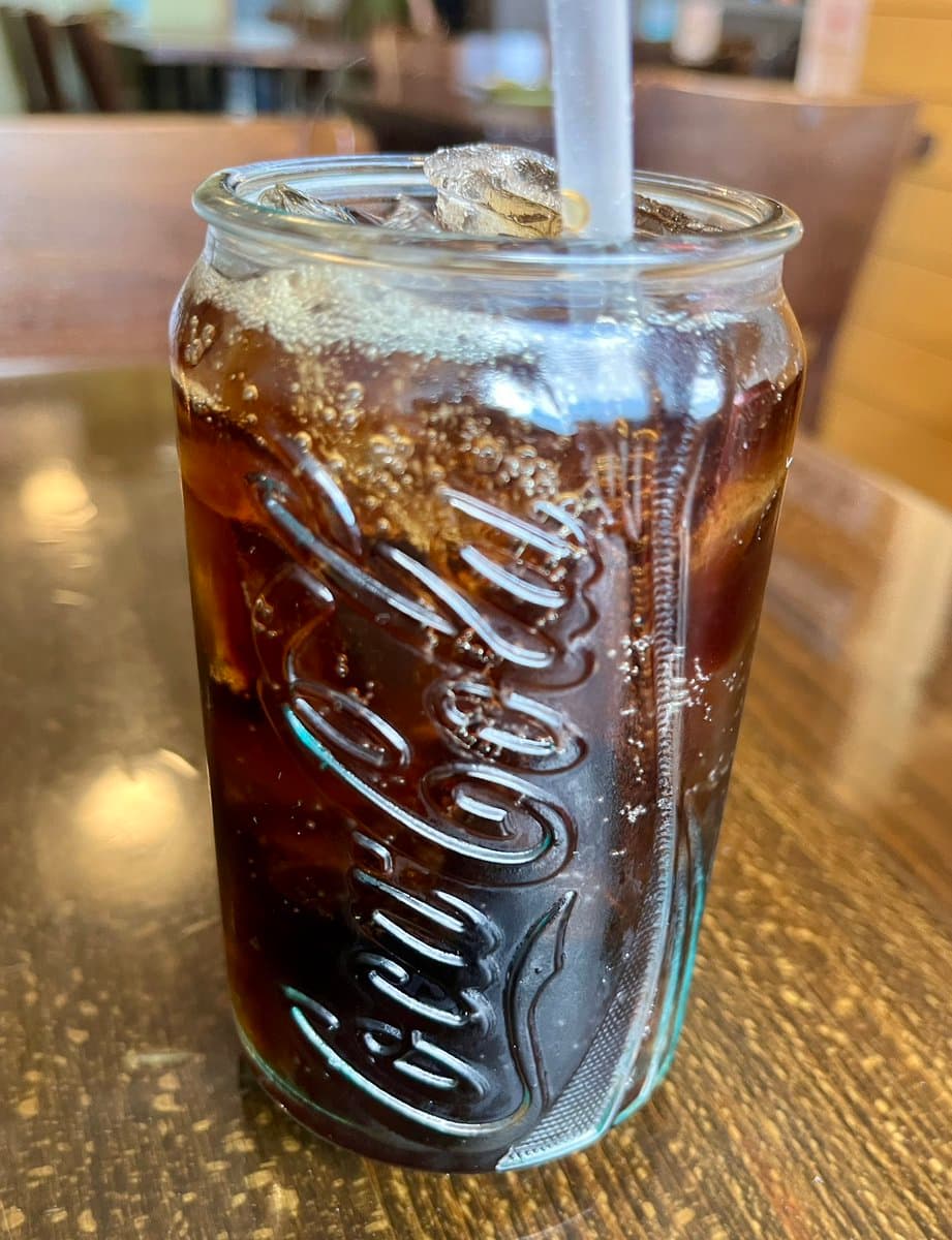A close-up photograph of a glass Coca-Cola bottle filled with a dark, carbonated beverage and ice, featuring a white straw. The bottle sits on a wooden table in what appears to be a restaurant or cafe setting.
